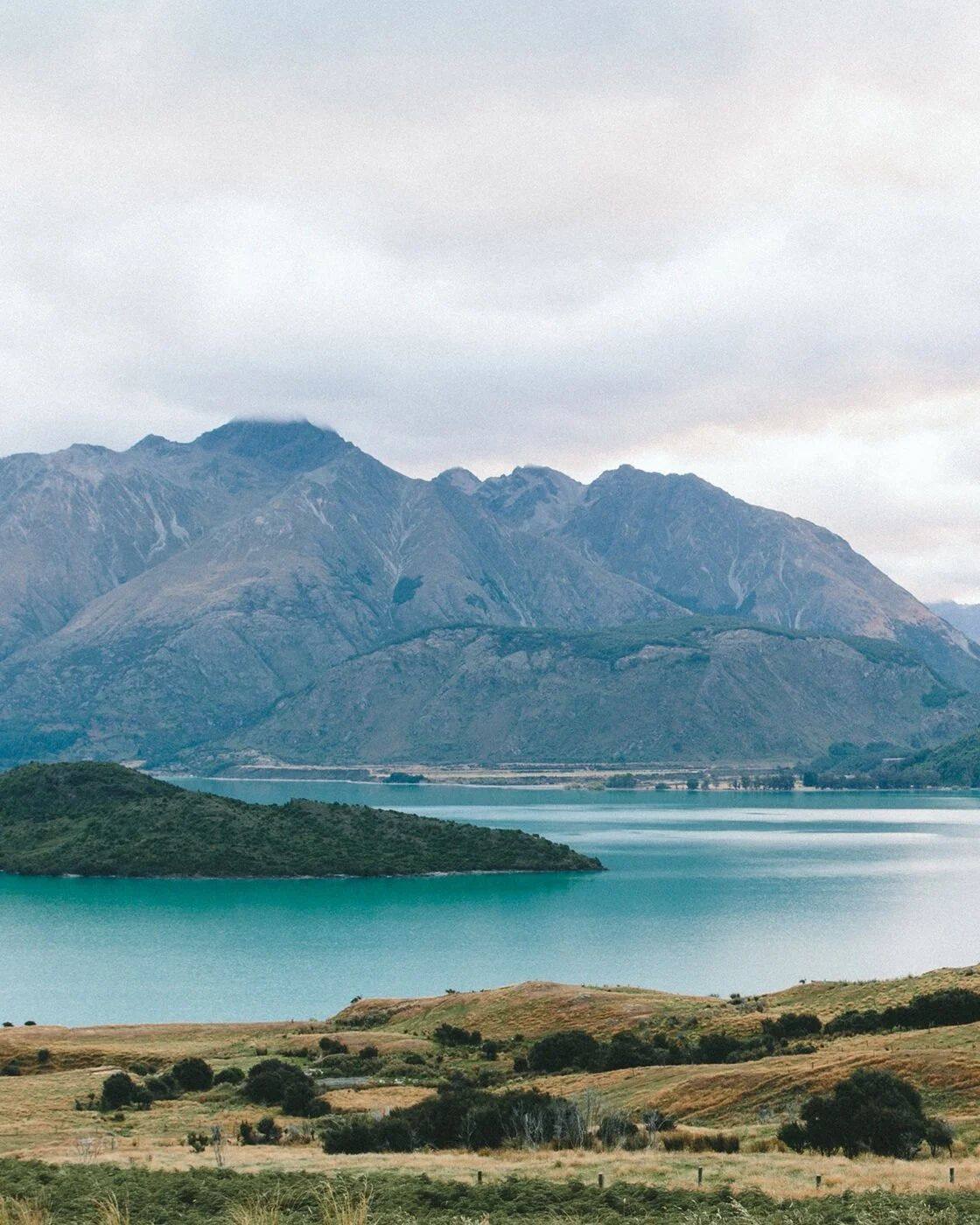 Scenic landscape of a mountain range with a lake in the foreground, under a cloudy sky.